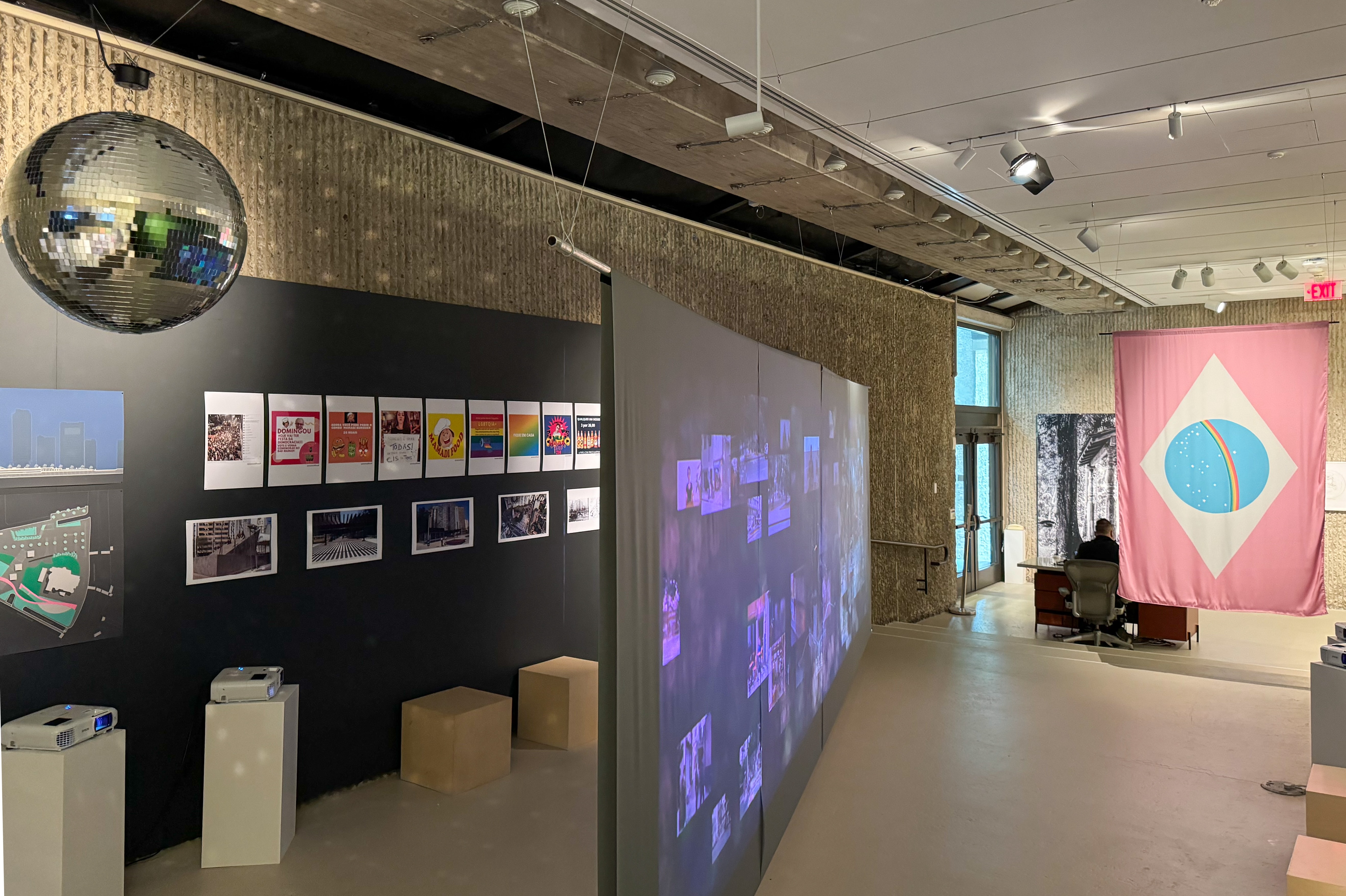 Another view of the exhibition showing a disco ball hanging from the ceiling on the left and a wall with several photos and posters, including colorful LGBTQ+ themed posters. A large video projection casts images across a screen positioned diagonally in the middle of the gallery. The pink flag with a blue circle and rainbow design is visible again in the background, hanging near an exit door. The room has a minimalist, concrete aesthetic, drawing attention to the images and projections.