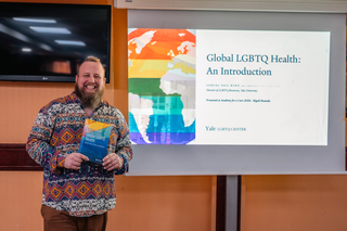 Director Samuel Byrd stands with Global LGBTQ Health book in front of title powerpoint slide titled "Global LGBTQ Health: An Introduction"