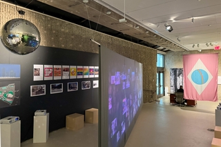 Another view of the exhibition showing a disco ball hanging from the ceiling on the left and a wall with several photos and posters, including colorful LGBTQ+ themed posters. A large video projection casts images across a screen positioned diagonally in the middle of the gallery. The pink flag with a blue circle and rainbow design is visible again in the background, hanging near an exit door. The room has a minimalist, concrete aesthetic, drawing attention to the images and projections.